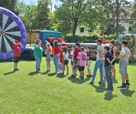 Eine Gruppe von Menschen steht auf einem Rasen, in der Nähe eines großen aufblasbaren Dartspiels und einer aufblasbaren Hindernisbahn, unter einem Zelt.