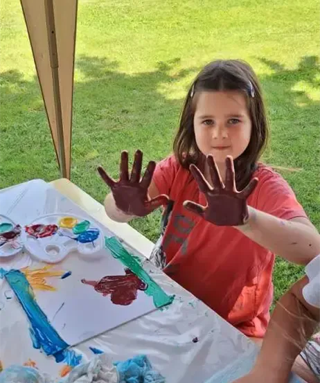 Ein kleines Mädchen zeigt ihre schokoladenverschmierten Hände nach dem Malen, sitzend an einem Tisch mit weißer Tischdecke und verschiedenen Malutensilien.