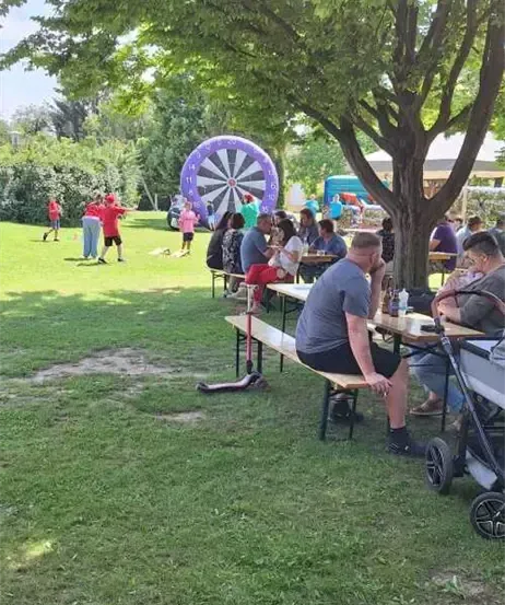 Menschen versammeln sich in einem Park unter einem Baum mit einem Dartboard, einige sitzen auf Bänken, während andere stehen. Ein Mann sitzt mit den Händen im Gesicht, und ein Kinderwagen ist in der Nähe.