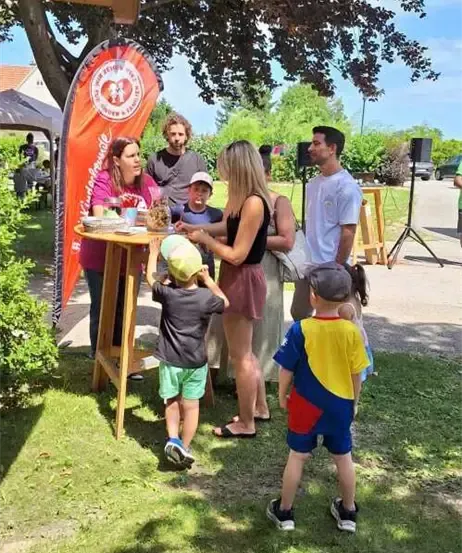 Eine Gruppe von Menschen, darunter Erwachsene und Kinder, versammelt sich um einen Tisch unter einem Zelt in einem Park. Eine Frau steht am Tisch mit verschiedenen Gegenständen, während ein Mann und ein Junge sich mit ihr beschäftigen.