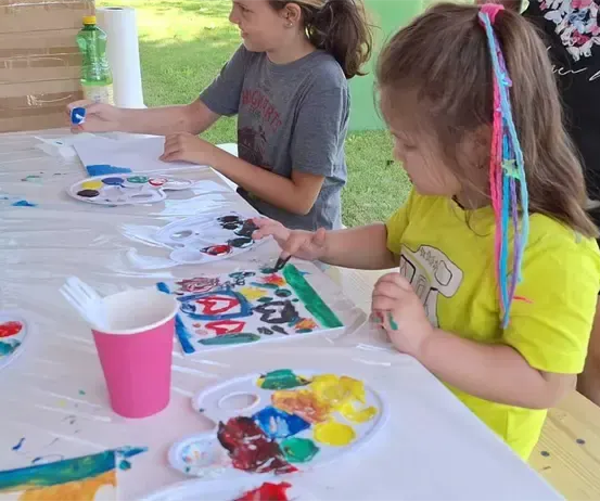 Zwei junge Mädchen malen auf einem Tisch mit bunter Farbe. Es gibt Teller mit Farbe, Becher und eine Holzbank hinter ihnen. Eine Person steht hinter dem Tisch.