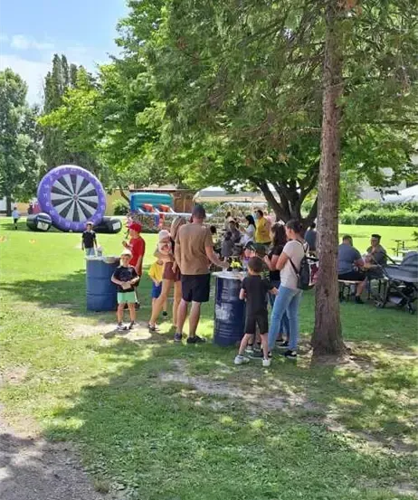 Eine Gruppe von Menschen, einschließlich Kindern, steht in einem Park mit Bäumen, Fässern und einem aufblasbaren Dartboard.