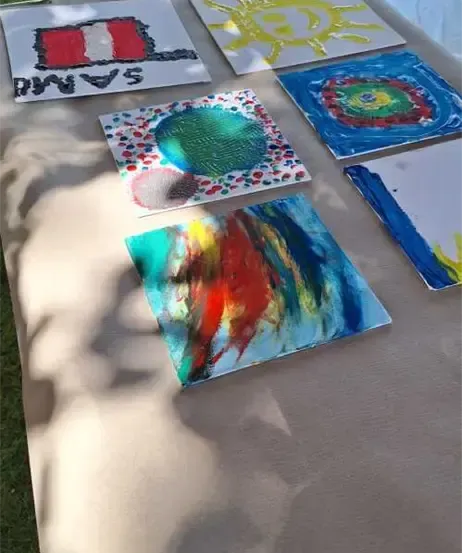 Mehrere Kunstwerke sind auf einem Tisch im Freien ausgestellt. Es gibt ein Flaggendesign, eine Sonne und farbenfrohe abstrakte Gemälde.