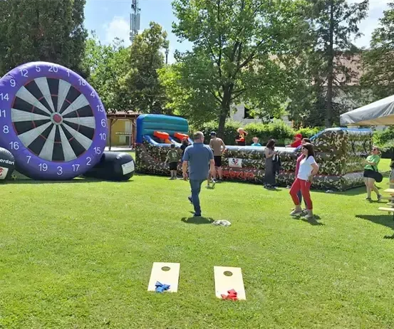Eine Gruppe von Menschen spielt im Park. Ein riesiger Dartautomat und aufblasbare Hindernisse sind aufgestellt. Ein Mann in einem blauen Shirt geht auf das Dartboard zu.