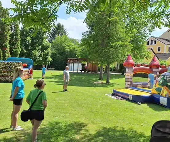 Zwei Frauen und ein Mann stehen in der Nähe von aufblasbaren Attraktionen in einem Park mit einem gelben Haus im Hintergrund.