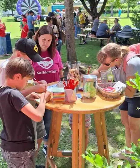 Zwei Kinder dekorieren Gegenstände auf einem Tisch in einem Park. Hinter ihnen schaut eine Frau in einem rosa T-Shirt mit einem Herz und dem Wort 'Solidaritat' zu ihnen hin. Es sind andere Leute um sie herum.