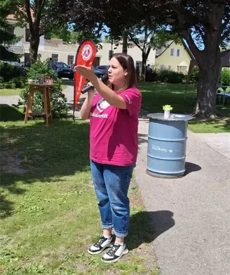 Eine Frau in einem rosa T-Shirt hält ein rotes Banner mit einem Mikrofon und spricht zur Menge.