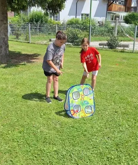 Zwei Kinder spielen mit einem bunten Pop-up-Spiel auf einem Rasen, mit einem weißen Haus und Zaun im Hintergrund.