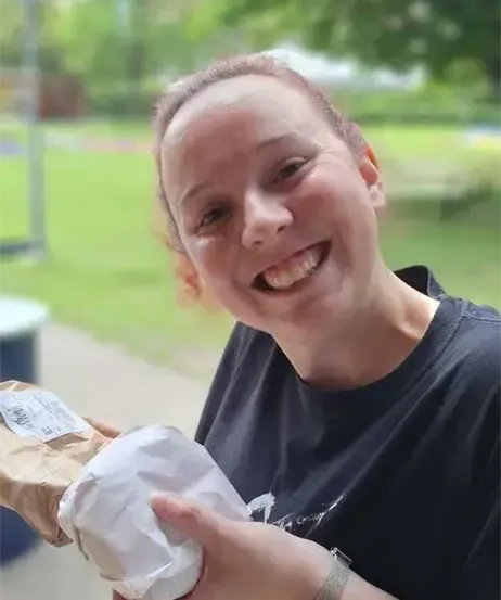 Eine Frau hält einen Papierbeutel und ein Sandwich draußen an einem sonnigen Tag. Sie trägt ein dunkles T-Shirt und eine silberne Uhr. Bäume und ein Feld sind im Hintergrund.
