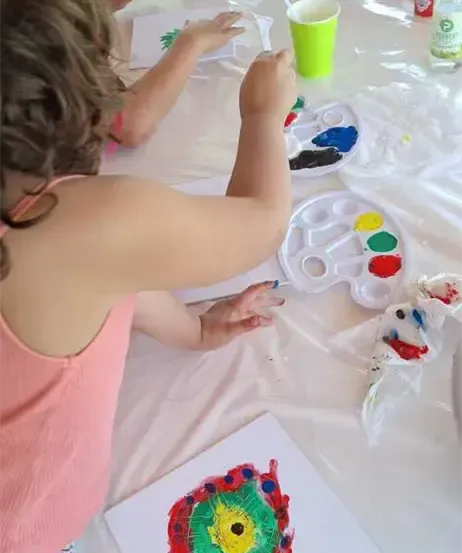 Zwei Kinder malen an einem Tisch mit weißem Tuch. Ein Kind malt einen bunten Kreis. Verschiedene Farben und Pinsel liegen auf dem Tisch.