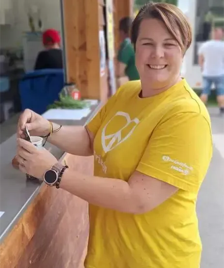 Eine Frau in einem gelben T-Shirt lächelt und hält einen Becher, während sie an einem Tresen steht. Hinter ihr steht eine Holzsäule.
