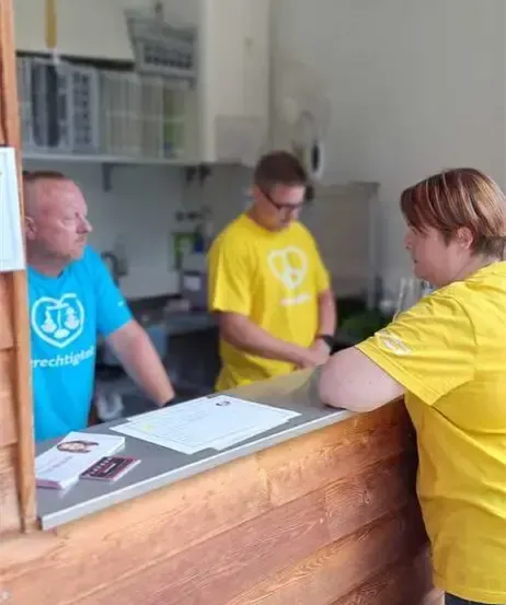 Drei Personen befinden sich in einem Raum mit einem Holztresen, einem Mann mit Brille und gelbem Shirt mit Logo und einer Frau in gelbem Shirt, die ihn ansieht.