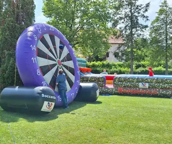 Eine Person steht vor einem großen aufblasbaren Dartboard auf einem Rasenfeld, mit anderen aufblasbaren Spielen in der Nähe. Dahinter stehen Bäume und ein Haus.