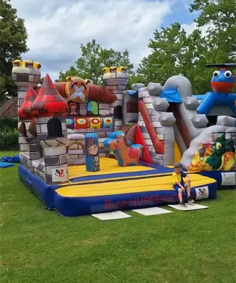 Ein aufblasbarer Sprungburg mit burgähnlicher Struktur, Rutschen und Figuren steht auf einem Rasenfeld mit Bäumen. Ein Junge sitzt in der Nähe auf dem Boden.