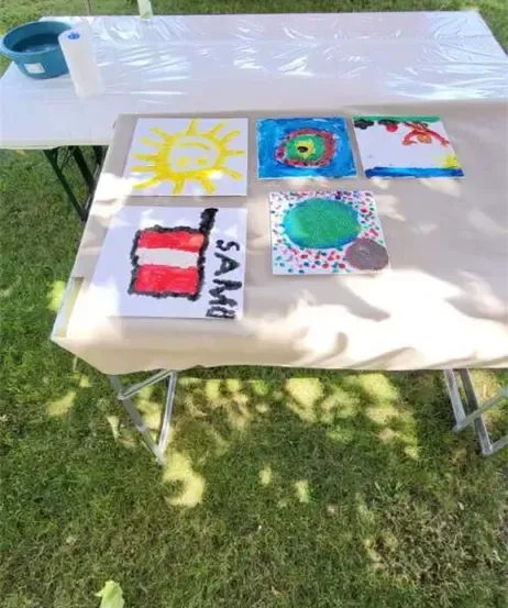 Ein Tisch mit einer weißen Abdeckung zeigt vier Gemälde. Zwei Bänke mit sitzenden Personen befinden sich hinter dem Tisch. Ein blauer Eimer steht auf der linken Seite.