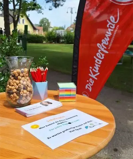 Ein Holztisch zeigt eine Schüssel mit Bohnen, bunte Stifte und eine Box mit Aufklebern. Es gibt ein Zertifikat, eine Flagge und ein Gebäude im Hintergrund.