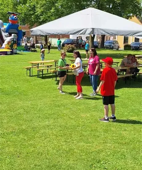 Eine Gruppe von Menschen versammelt sich in einem Park. Einige sitzen an Picknicktischen unter einem Zelt. Ein Junge mit rotem Hut spielt mit einem Ball. In der Nähe gibt es aufblasbare Spielgeräte und geparkte Autos.