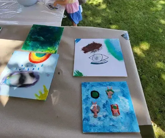 Ein Tisch, der mit Kunstwerken bedeckt ist, darunter ein Gemälde mit einem Regenbogen, einem Auge und drei Personen im Wasser. Ein Kind steht in der Nähe.