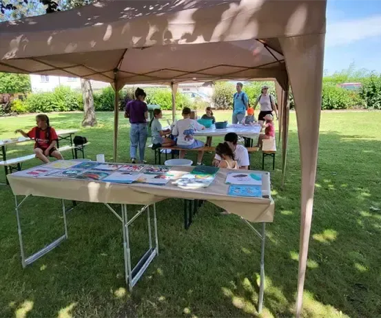 Eine Gruppe von Kindern bastelt unter einem Zelt in einem Park. Tische mit Papieren und Zubehör sind aufgestellt.