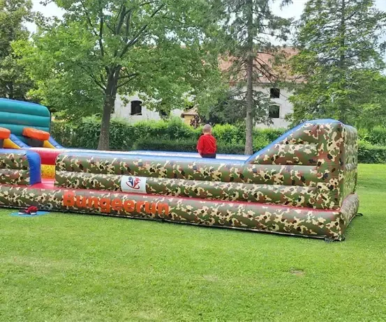 Ein junger Junge steht in der Nähe eines großen, aufblasbaren Hindernisparcours mit Tarnmuster auf einer Rasenfläche. Bäume und ein Haus sind im Hintergrund.