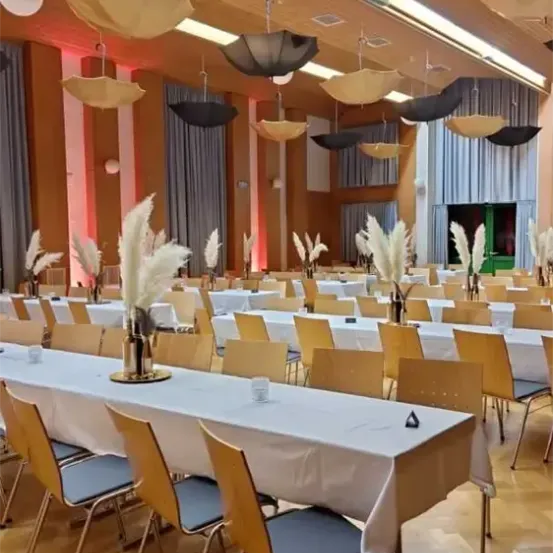 Ein Bankettsaal ist mit langen Tischen eingerichtet, die mit weißen Tischdecken bedeckt sind. Die Stühle sind um die Tische herum angeordnet, und dekorative Regenschirme hängen von der Decke. Der Raum ist mit warmer Beleuchtung erhellt.