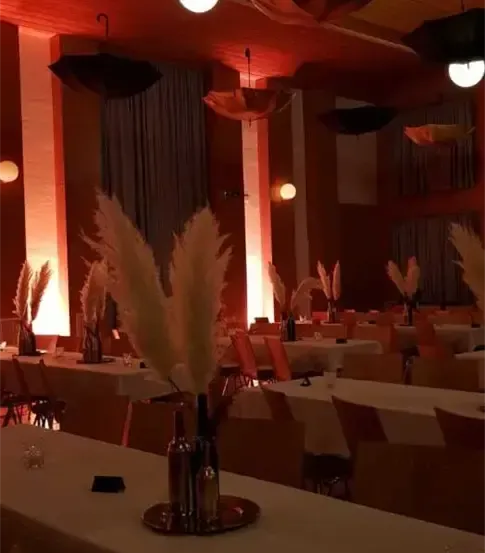 Ein leerer Bankettsaal mit Tischen, Stühlen und dekorativen Elementen. Die Tische haben weiße Tischdecken, und es gibt große Vasen mit Pampasgras.