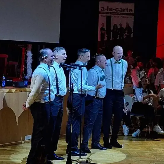 Eine Gruppe von Männern in Anzügen und Krawatten tritt mit Mikrofonen vor einem Publikum auf der Bühne auf.