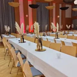 Ein Raum mit langen Tischen und Stühlen, in Weiß gehüllt, für eine Veranstaltung vorbereitet. Topfpflanzen und Regenschirme schmücken die Tische. Rote Beleuchtung und hängende Regenschirme verleihen dem Raum eine festliche Atmosphäre.