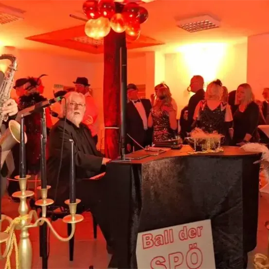 Ein Mann spielt Saxophon, während ein anderer Mann Klavier spielt bei einer festlichen Veranstaltung mit einer Menschenmenge, roten Luftballons und einem Banner, das 'Ball der SPO' sagt.