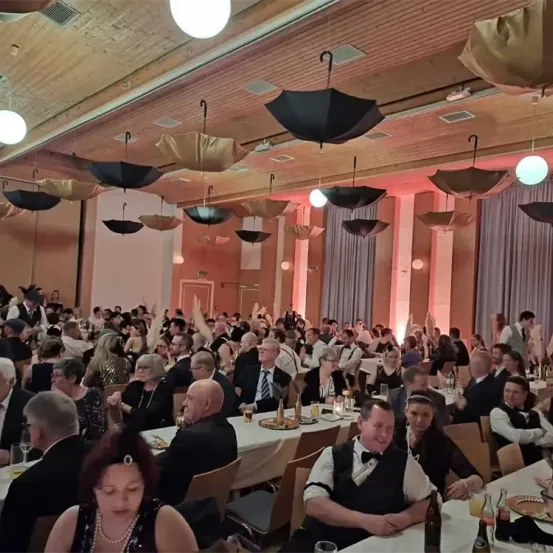 Ein großer Saal mit vielen Menschen in formeller Kleidung. Tische mit Tellern, Weingläsern und Flaschen. Die Leute klatschen und heben die Hände. Mehrere Regenschirme hängen von der Decke.