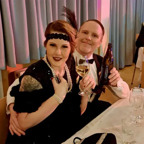 Eine Frau und ein Mann in Vintage-Kleidung, die eine Flasche und ein Weinglas halten, lächeln und posieren für ein Foto. Hinter ihnen sind Vorhänge, und es gibt zusätzliche Weingläser und eine Tasche auf dem Tisch.