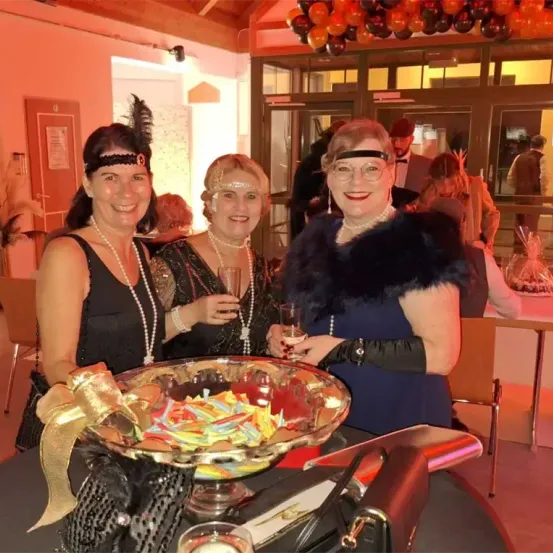 Drei Frauen in 1920er-Jahre-Kleidung stehen an einem Tisch mit einer Schüssel voller Süßigkeiten und lächeln für ein Foto. Dahinter findet eine Party statt, mit Luftballons und einer festlichen Atmosphäre.