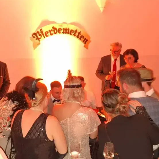 Eine Gruppe formell gekleideter Personen versammelt sich um einen Tisch, einige halten Gläser mit Wein in der Hand. Der Raum ist von einem gelben Licht beleuchtet, und über ihnen ist ein Schild mit der Aufschrift 'Verbeugen' zu sehen.