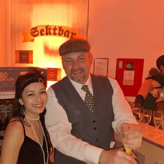 Ein Mann und eine Frau in 1920er-Kleidung lächeln und halten eine Flasche in einer Bar, mit einem Schild über ihnen, das 'Gektbar' steht.
