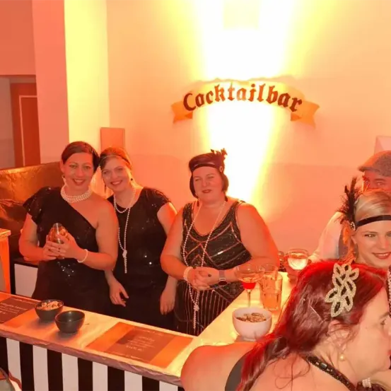 Mehrere Frauen in 1920er-Kleidung stehen an einer Cocktailbar, halten Getränke und lächeln. Dahinter steht ein Schild mit der Aufschrift Cocktailbar. Ein Mann mit Hut sitzt in der Nähe.