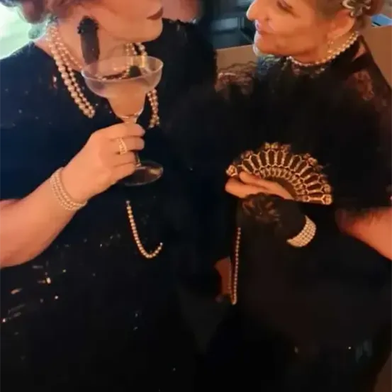 Zwei Frauen in schwarzen Vintage-Flapper-Kleidern, die einen Cocktail und einen Fächer halten, stehen möglicherweise auf einer Party eng beieinander.