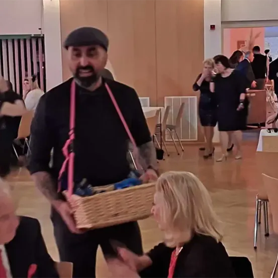 Ein Mann in einem schwarzen Shirt und einer Mütze trägt einen Korb, während eine Frau in einem schwarzen Kleid zusieht. Andere Menschen im Raum unterhalten sich.