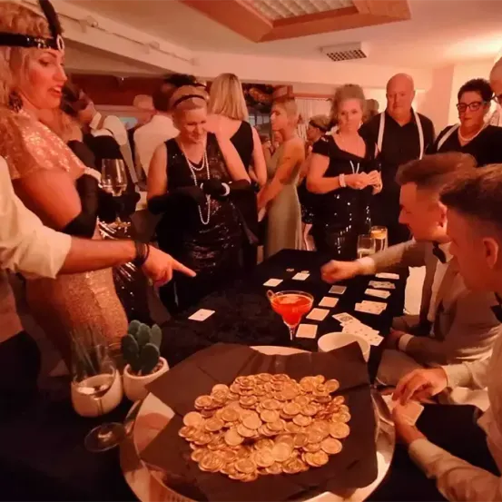 Eine Gruppe von Menschen, die in 1920er-Jahre-Kleidung gekleidet sind, versammelt sich um einen Tisch mit Spielaufbau. Eine Frau zeigt auf den Tisch, und andere sitzen oder stehen in der Nähe.