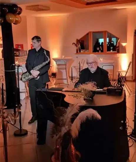 Ein Musiker spielt Saxophon, während ein anderer am Klavier sitzt in einem dekorierten Raum mit Luftballons und warmer Beleuchtung.