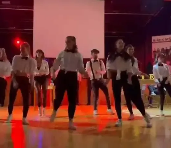 Eine Gruppe von Menschen tanzt auf einer Bühne mit roter Beleuchtung. Sie tragen weiße Hemden und schwarze Hosen.