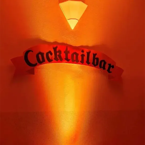 Eine Wand mit einer Lampe und dem Wort 'Cocktailbar' in Schwarz, mit orangefarbenem Licht, das sich an der Wand spiegelt.