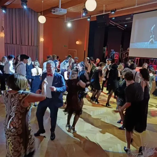 Eine Gruppe von Menschen tanzt auf einem Holzboden in einem Saal. Sie tragen elegante Kleider. Eine Projektorlampe ist an der Decke montiert.