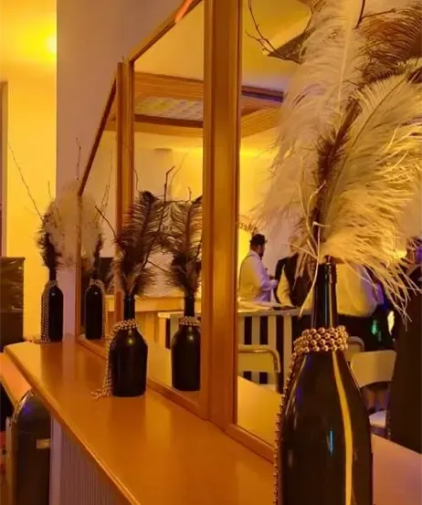 Ein hölzerner Tresen mit schwarzen Vasen, die Federn und Ketten halten. Dahinter reflektiert ein Spiegel einen Raum mit einem Tisch, Stühlen und Menschen.