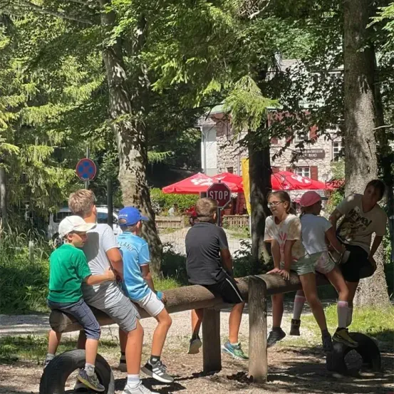 Mehrere Kinder sitzen auf einer Holzbalken in einem Park mit grünen Bäumen und einem Stoppschild in der Nähe. Dahinter befindet sich ein Gebäude mit roten Sonnenschirmen.