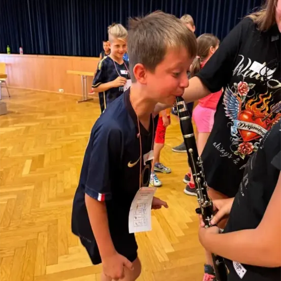Mehrere Kinder spielen Instrumente in einem Raum. Ein Junge spielt Klarinette und ein Mädchen Gitarre. Sie alle lächeln.