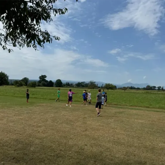 Eine Gruppe von Menschen spielt ein Spiel auf einem Rasenfeld unter einem blauen Himmel mit verstreuten Wolken. Einige tragen Sporttrikots.