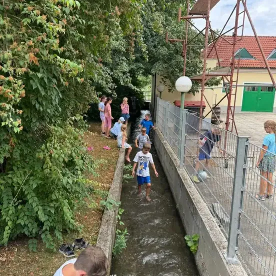 Kinder spielen in einem flachen Bach in der Nähe eines Hauses mit rotem Dach und grünen Türen. Bäume und Pflanzen umgeben das Gebiet.