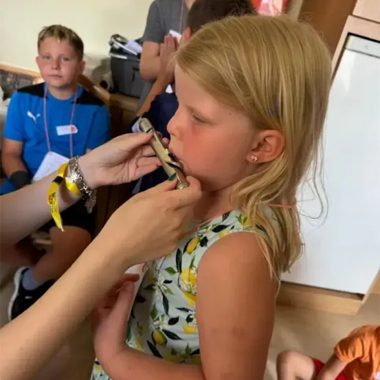 Ein junges Mädchen spielt eine Flöte, während eine andere Person sie für sie hält. Andere Kinder sitzen in der Nähe, eines trägt ein blaues Shirt und Turnschuhe.