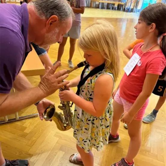 Ein Mann hilft einem kleinen Mädchen, ein Saxophon zu spielen, während andere in einer Halle beobachten.