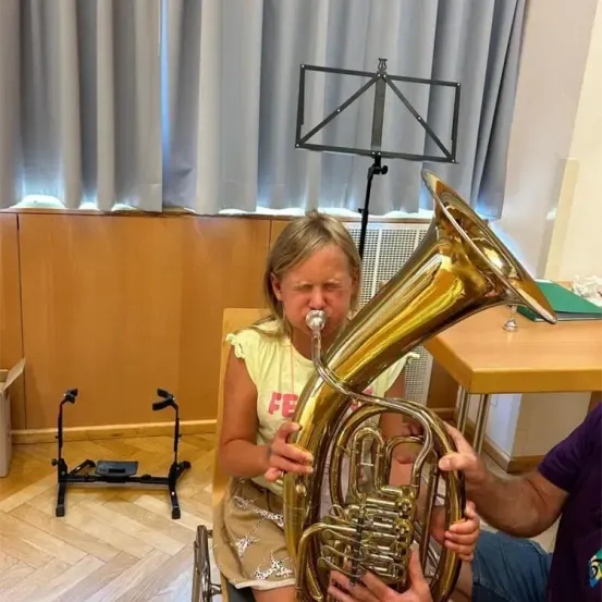 Ein junges Mädchen spielt ein großes Blechblasinstrument, möglicherweise eine Tuba, während sie auf einem Stuhl sitzt. Ein Mann hilft ihr dabei. Dahinter befindet sich eine Holzwand mit einem Notenständer und Vorhängen.